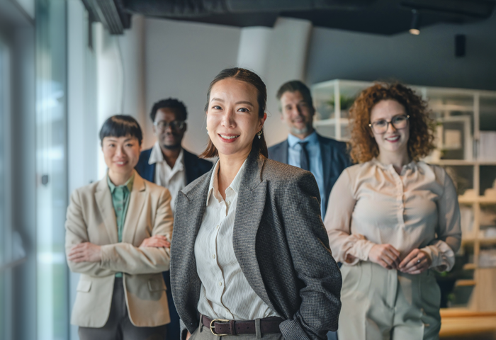 A diverse group of five professionals stands together in a modern office. A smiling woman in a gray blazer stands confidently in the foreground with her hands in her pockets, while four colleagues stand behind her, smiling toward the camera.